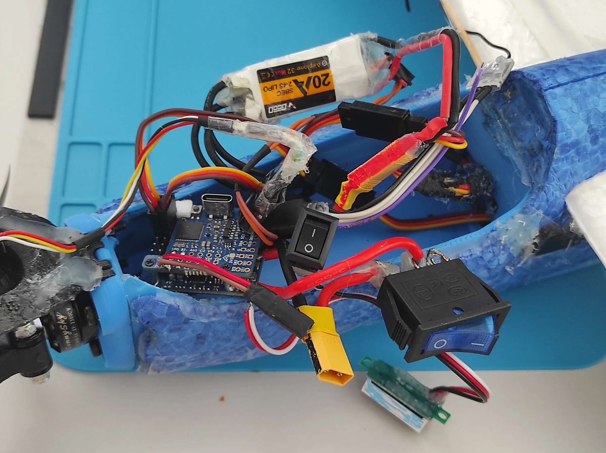 an opened blue fuselage with a flight controller, a white ESC labelled 20A, two rocker switches, a yellow connector and a small voltmeter board