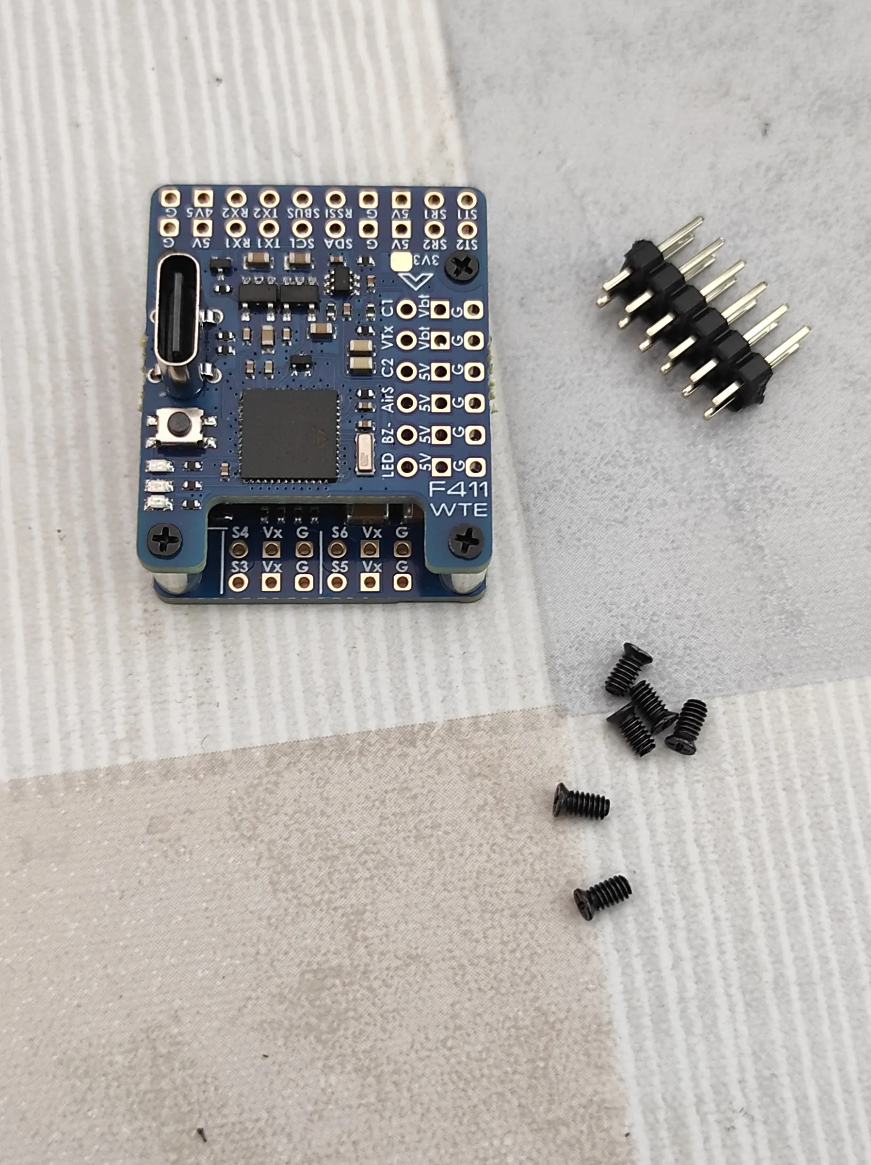 a blue Matek F411-WTE flight controller with no wires, a header strip and screws beside it